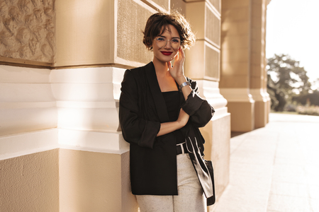 Joyful lady in jacket and white pants smiling outside. Lovely woman with bright lips in eyeglasses looking into camera outdoors.